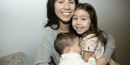 A mother holds her two small children in her arms, smiling.