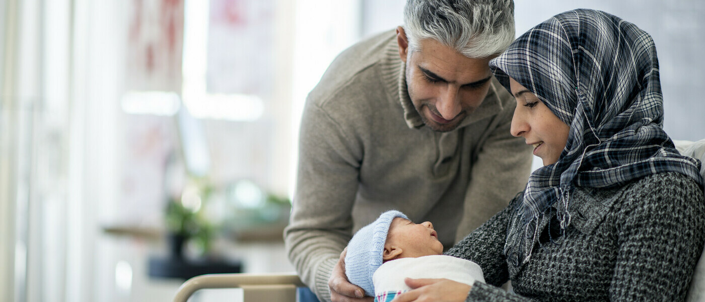 A woman sits holding a baby. A man leans over and rests his hand on the back of the baby's head.